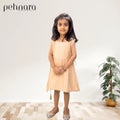 Young girl in a beige dress standing in a room with a plant and 'pehnara' branding.