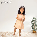 Young girl wearing a beige dress standing in a room with a plant and 'pehnara' branding.