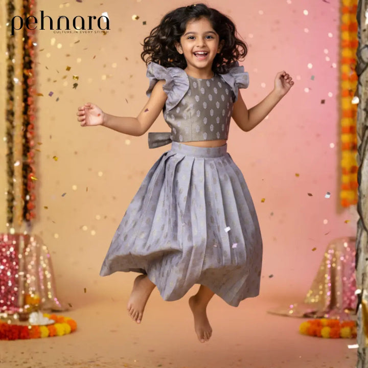 Child in a gray dress with ruffled sleeves and a pleated skirt, jumping in front of a decorative background with lights and confetti.
