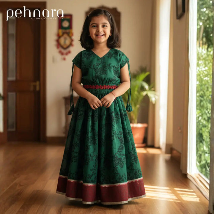 Young girl in a green traditional dress standing in a well-lit room with a window in the background.