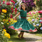 Young girl in a green dress with red trim standing in a garden with colorful flowers.